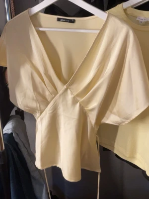 Ljusgul omlottopp från Gina Tricot - Supersnygg ljusgul omlottopp från Gina Tricot med korta, vida ärmar och knytband i midjan. Toppen har djup v-ringning och ett mjukt, glansigt tyg som ger en elegant känsla. Perfekt att styla med jeans eller kjol för en trendig look.