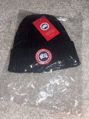 Svart ribbstickad mössa Canada Goose - Svart ribbstickad mössa från Canada Goose med klassisk rund logotyp framtill. Mössan har uppvikt kant och är tillverkad i ett mjukt material som håller dig varm. Perfekt för kalla dagar och snygg att matcha med vinterjackan.