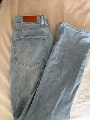 Ljusblå raka jeans i denim - Säljer ett par oanvända ljusblå jeans med rak passform och klassisk femficksdesign. Jeansen har en snygg läderpatch i midjan. Tillverkade i ett slitstarkt bomullsmaterial som ger en avslappnad look.