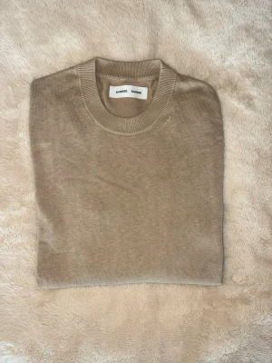 Beige stickad tröja från Samsoe Samsoe - En stilren beige stickad tröja från Samsoe Samsoe med rund halsringning och långa ärmar. Tröjan har ribbade muddar vid ärmslut och nederkant. Perfekt basplagg i mjukt material som passar till allt.