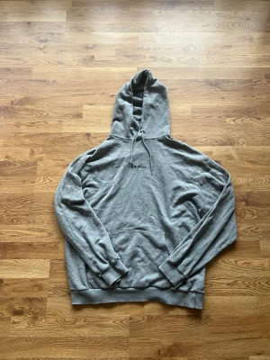 Grå hoodie med broderad text, Kolla alla bilder!😌 - Säljer en stilren grå hoodie med huva och dragsko. Framtill finns en broderad text och en klassisk magficka. Hoodien har ribbade muddar vid ärmslut och nederkant. Perfekt för en avslappnad och trendig look.