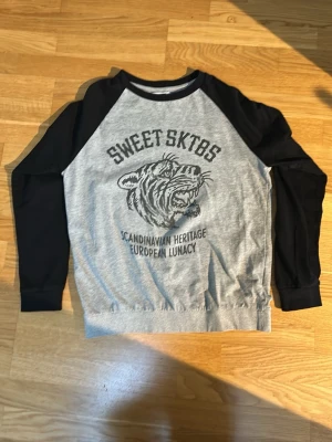 Sweet SKTBS grå och svart hoodie - Grå hoodie från Sweet SKTBS med svarta ärmar och tryck av en tiger på bröstet. Texten 'Scandinavian Heritage European Lunacy' under motivet. Klassisk raglanmodell med rund hals. Perfekt för en streetwear-look.