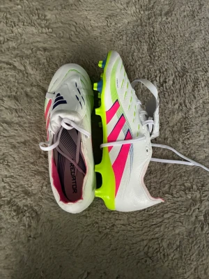 Adidas Predator vita fotbollsskor - Säljer ett par Adidas Predator fotbollsskor i vitt med rosa och mörkblå detaljer samt neongul sula. Skorna har klassisk snörning och är tillverkade i syntetmaterial för bra grepp och kontroll på planen. Perfekta för dig som vill sticka ut på matchen!
