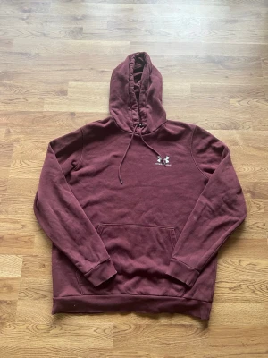 Vinröd hoodie från Under Armour - Säljer en vinröd hoodie från Under Armour med klassisk känguruficka och justerbar huva med dragsko. Tröjan har ribbade muddar vid ärmslut och nederkant samt en liten vit logga på bröstet. Perfekt för en avslappnad och sportig stil.