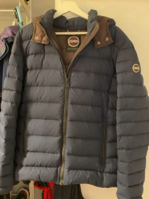 Blå pufferjacka från Colmar - Snygg blå pufferjacka från Colmar med huva och bruna detaljer vid kragen. Jackan har två sidofickor med dragkedja, Colmar-logga på ärmen och quiltad design. Perfekt för kalla vinterdagar och riktigt skön att bära. Tveka inte på att höra av sig vid frågor😁