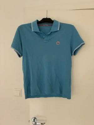 Blå pikétröja från Moncler - Snygg blå pikétröja från Moncler med klassisk krage och knappar framtill. Tröjan har vita detaljer på krage och ärmslut samt Moncler-logga på bröstet. Perfekt till sommaren och enkel att matcha med jeans eller shorts.