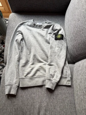 Grå sweatshirt från Stone Island - Snygg grå sweatshirt från Stone Island med klassisk rund hals och ribbade muddar. Tröjan har den ikoniska svarta patchen med gul och grön logga på vänster ärm. Perfekt för en clean och stilren look.