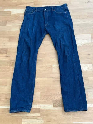 Levi's 501 raka blå jeans - Klassiska Levi's 501 jeans i mörkblå denim med raka ben och fem fickor. Jeansen har den ikoniska läderpatchen bak i midjan och röd Levi's-tagg på bakfickan. Perfekta för en tidlös och avslappnad stil.