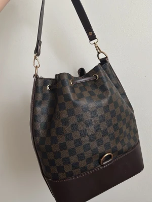 Louis Vuitton brun bucket bag - Säljer en brun bucket bag i klassiskt Damier-mönster. Väskan har justerbar axelrem, dragskostängning med guldfärgade detaljer och mörkbruna läderdetaljer. Perfekt för dig som gillar lyxig och tidlös design.
