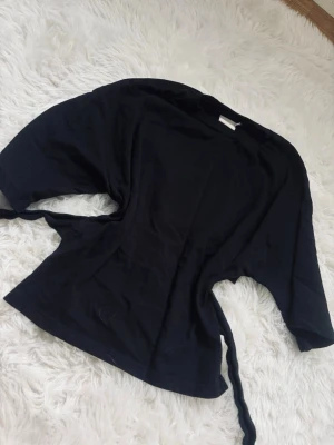 svart  omlottopp med knytband - Stilren svart omlottopp med knytband i midjan och trekvartsärm. Perfekt för dig som gillar en enkel men trendig look. Tillverkad i mjukt tyg som känns skönt mot huden.