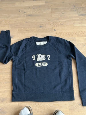 Mörkblå sweatshirt Abercrombie & Fitch - Mörkblå sweatshirt med broderad bulldog, siffrorna 9 och 2 samt texten A&F på bröstet. Tröjan har rund halsringning och långa ärmar. Perfekt för en avslappnad och sportig stil.