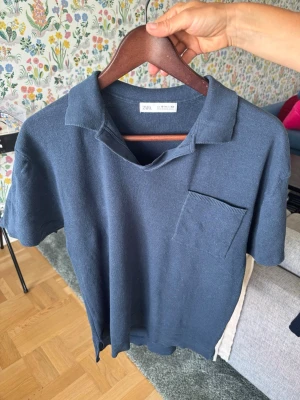 Mörkblå stickad polotopp från Zara - Mörkblå stickad polotopp från Zara med korta ärmar och en bröstficka. Toppen har en klassisk krage och är tillverkad i ett mjukt stickat material som ger en avslappnad men ändå stilren look.