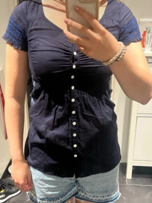 Navy Blue Button-Up Top with Embroidery - This is a navy blue short-sleeve blouse featuring white buttons down the front and a ruched bust. The sleeves have a blue crochet or lace detail. The second image shows a close-up of intricate floral embroidery in a lighter blue thread, likely on the same top. The blouse is paired with light blue denim shorts with a frayed hem in the first image. The overall style is casual and feminine, suitable for everyday wear.