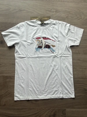 Vit t-shirt med broderad detalj Burberry - Vit t-shirt från Burberry med en broderad färgglad figur på bröstet. Klassisk passform med korta ärmar och rund halsringning. Tillverkad i 100% bomull för en mjuk och bekväm känsla.