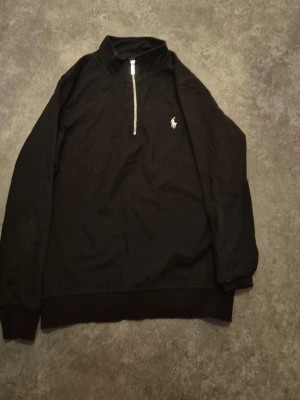 Svart half-zip hoodie från Polo Ralph Lauren - Svart hoodie med halv dragkedja och hög krage från Polo Ralph Lauren. Klassisk design med vit broderad logga på bröstet. Mjuk bomullskänsla och långa ärmar, perfekt för en avslappnad och stilren look.
