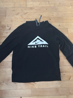 Svart Nike Trail hoodie med tryck - Säljer en svart hoodie från Nike Trail med stort vitt tryck på bröstet och justerbar huva med svartvita snören. Hoodien har en klassisk passform och är tillverkad i mjukt material som passar perfekt till chill eller träning.