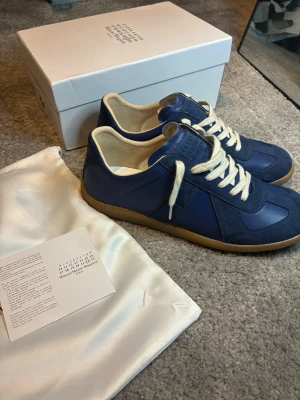 Blå sneakers från Maison Margiela - Snygga blå sneakers från Maison Margiela med beige sula och vita skosnören. Skorna har en mix av mocka och läderdetaljer, samt klassisk låg profil och rundad tå. Perfekta för dig som vill ha en stilren och trendig look.