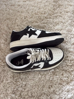 Svart och vita Bape Sta sneakers - Säljer ett par svarta och vita Bape Sta sneakers med klassisk stjärndesign på sidan. Skorna har lågt skaft, vita snören och svart yttersula. Ovandelen är i blankt läder med perforerad tåbox för extra ventilation. Perfekta för dig som gillar streetwear. Använda max 10 gånger, inte rört på 3 år och vill bli av med dom. Storlek 43. Skriv om du har frågor om skorna eller pris, är ganska flexibel 