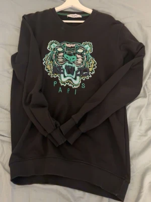 Svart sweatshirt med tigertryck Kenzo - Svart sweatshirt från Kenzo med stort broderat tigerhuvud i turkos, vitt och grönt på bröstet samt texten 'KENZO PARIS' under motivet. Rund halsringning och långa ärmar. Tillverkad i mjukt bomullsmaterial för en skön känsla.