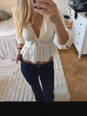 Vit broderad blus med v-ringning - Söt vit blus med broderade detaljer och djup v-ringning. Blusen har trekvartsärm och volang nedtill som ger en feminin siluett. Perfekt till jeans eller kjol för en trendig look. Tillverkad i ett lätt och luftigt tyg.