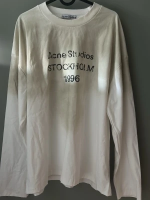 Vit långärmad Acne Studios t-shirt - Vit långärmad t-shirt från Acne Studios med tryck i svart text på bröstet där det står 'Acne Studios STOCKHOLM 1996'. T-shirten har en klassisk rund halsringning och är gjord i mjuk bomull. Perfekt för en clean och stilren look.