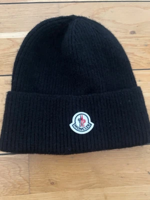 Svart ribbstickad Moncler mössa - Säljer en svart ribbstickad mössa från Moncler med uppvikt kant och Moncler-logga framtill. Klassisk och stilren design som passar perfekt till kalla dagar. Mössan är mjuk och sitter skönt på huvudet.