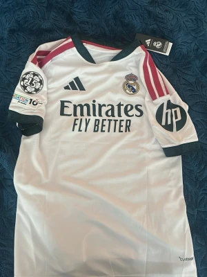 Real Madrid Mbappé matchtröja - Säljer en vit Real Madrid matchtröja från Adidas med Mbappé och nummer 10 på ryggen. Tröjan har svarta detaljer, röda ränder på axlarna, klubbmärke och sponsorloggor på bröstet samt HP-logga på ärmen. Tillverkad i lätt funktionsmaterial, perfekt för fotboll.