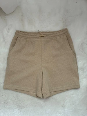 Beige shorts med snörning och fickor - Säljer ett par beige shorts med diskret sicksackmönster. De har elastisk midja med snörning och två sidofickor. Materialet känns mjukt och bekvämt, perfekt för varma dagar. Passformen är normal och shortsen har en enkel, clean look. Pris kan diskuteras vid snabb affär!
