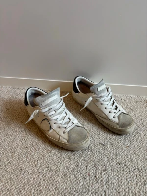 Philippe Model - Säljer ett par vita sneakers från Philippe Model med svart hälflik och grå detaljer på tån. Skorna har klassisk snörning, perforerade sidor och är tillverkade i läder med mockadetaljer. Perfekta för en clean och stilren look. Priset kan diskuteras!😉😃