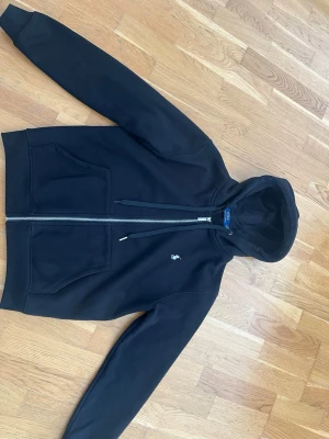 Polo Ralph Lauren Zip Hoodie Svart  - Säljer en Polo Ralph Lauren Zip Hoodie i svarta som är i mycket bra skick. Tröjan har bara använts 2-3 gånger då den blev liten på mig. Tröjan är köpt för ungefär 1500.