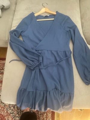 Blå omlottklänning med volang - Snygg blå kortklänning med omlottmodell och knytband i midjan. Klänningen har långa ärmar och volang nedtill för en extra feminin touch. Perfekt för dig som gillar en avslappnad men ändå chic stil.
