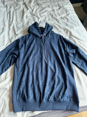 Mörkblå hoodie med mönster - Snygg mörkblå hoodie med huva och dragsko. Hoodien har ett diskret, upprepat mönster i tyget som ger en exklusiv känsla. Ribbstickade muddar vid ärmslut och nederkant. Perfekt för dig som vill ha en stilren men ändå unik look.