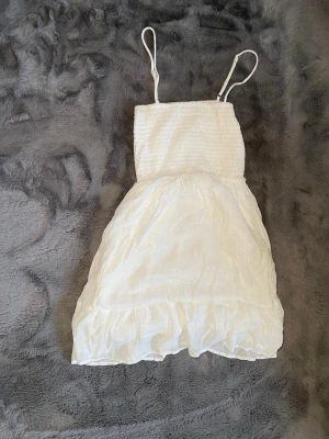 Vit sommarklänning från FB Sister - Söt vit klänning från FB Sister i storlek XS. Klänningen har smala axelband, smockad överdel och volangkant nedtill. Perfekt för varma dagar och har en lätt, luftig känsla. Materialet är troligtvis bomull eller polyester.