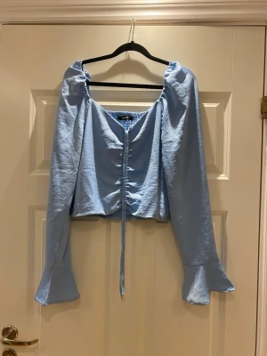 Ljusblå blus med puffärm och snörning - Trendig ljusblå blus med långa puffärmar och volang vid ärmslut. Blusen har en djup urringning och snörning framtill som ger en snygg detalj. Perfekt för dig som vill ha en feminin och modern look.