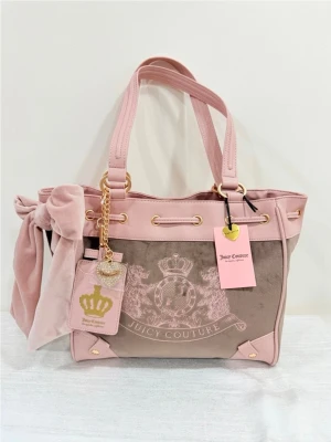 Rosa handväska Juicy Couture - Snygg rosa handväska från Juicy Couture i mjuk velour och detaljer i guld. Väskan har ett stort rosettband på sidan, guldfärgade metalldetaljer, och ett emblem med krona och logga framtill. Två axelband och dekorativ tag med hjärta och krona.