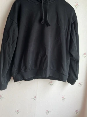 Svart hoodie med huva och dragsko - En klassisk svart oversized hoodie med huva och dragsko. Hoodien har långa ärmar, ribbade muddar och en avslappnad passform. Tillverkad i mjukt sweatshirtmaterial som är perfekt för chill dagar. Enkel och stilren design utan tryck eller logga.