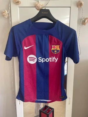 Barcelona matchtröja Nike blå/röd - Säljer en officiell FC Barcelona matchtröja från Nike i blått och rött med klubbmärke, Nike-logga och Spotify som sponsor framtill. Tröjan har korta ärmar, sportig passform och UNHCR-logga på ryggen. Perfekt för fotbollsfans!