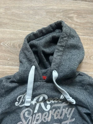 Grå hoodie med tryck och huva - Mysig grå hoodie med stor huva och vita dragsnören. Framsidan har ett stort, stilrent tryck i vitt med texten 'The Real Superdry'. Perfekt för en avslappnad och trendig look. Tillverkad i mjukt material för extra komfort.