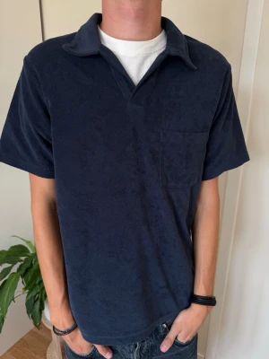 Mörkblå frotté polotopp - Mörkblå polotopp i mjuk frotté med korta ärmar och bröstficka. Avslappnad passform och klassisk krage utan knappar. Perfekt för en chill och stilren look.