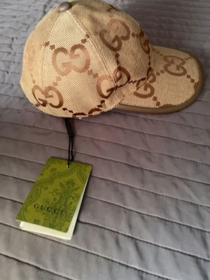 Beige keps från Gucci med GG-mönster - Snygg beige keps från Gucci med det klassiska GG-monogrammet i brunt över hela kepsen. Kepsen har böjd skärm och justerbar rem bak. Tillverkad i canvasmaterial med detaljer i brunt. Perfekt för dig som vill ha en exklusiv och trendig accessoar.