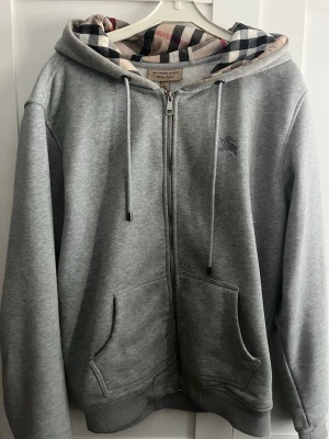 Grå hoodie med Burberry-rutor - Snygg grå zip-hoodie från Burberry med klassiskt rutigt foder i huvan. Hoodien har dragsko i huvan, två fickor fram och ribbade muddar. Perfekt för dig som vill ha en stilren men ändå lyxig look. Materialet känns mjukt och bekvämt.