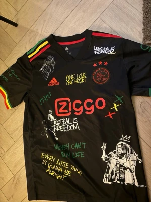 Ajax specialtröja – Ny -   Säljer en Ajax-tröja i svart med färgglada detaljer och unikt Bob Marley-inspirerat motiv. Bekväm passform och perfekt för både fotboll 