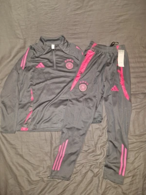 Grått träningsset Adidas Tyskland - Snyggt träningsset från Adidas med Tysklands landslagslogga. Setet är grått med rosa detaljer och består av en långärmad tröja med halv dragkedja och matchande byxor. Tillverkat i ett lätt och ventilerande material, perfekt för träning eller uppvärmning.