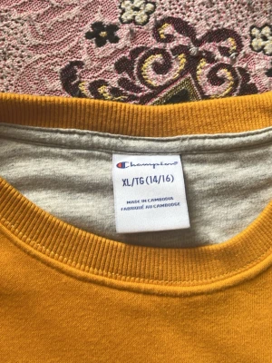 Gul stickad tröja från Champion - Gul stickad tröja från Champion i storlek XL. Tröjan har rund halsringning, långa ärmar och diskret Champion-logga på bröstet samt vid ärmslutet. Mjuk insida och ribbade muddar för extra komfort. Perfekt för dig som gillar streetwear och enkel stil.
