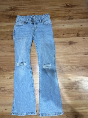 Ljusblå bootcut jeans med slitningar - Säljer ett par ljusblå bootcut jeans med slitningar över båda knäna. Jeansen har klassisk femficksmodell, normal midja och är tillverkade i denim. Perfekta för dig som gillar en avslappnad och trendig stil.