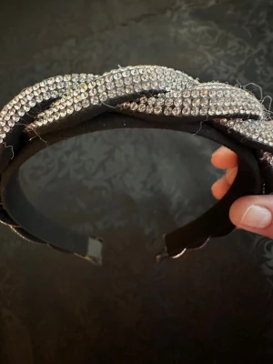 Flätat diadem med strass - Snyggt svart diadem med flätad design täckt av glittrande strass. Perfekt för dig som vill ha en extra touch av bling till din look. Diademet har en stadig form och gnistrar i ljuset. Materialet är textil och plast med fastlimmade stenar.