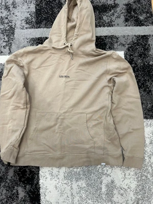 Beige hoodie från Les Deux - Snygg beige hoodie från Les Deux med broderad logga på bröstet. Klassisk känguruficka framtill och justerbar huva med dragsko. Tillverkad i mjukt bomullsmaterial som är skönt att bära. Perfekt för en avslappnad och trendig look.