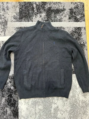 Mörkgrå ziptröja från Polo Ralph Lauren - Mörkgrå stickad ziptröja från Polo Ralph Lauren med hög krage och två fickor framtill. Tröjan har diskret blå logga broderad på bröstet och ribbade muddar vid ärmslut och nederkant. Perfekt för lager på lager och stilren look.
