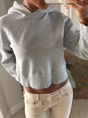 Grå croppad hoodie - Säljer en ljusgrå croppad hoodie med huva och ribbade muddar vid ärmslut och nederkant. Hoodien har en enkel och clean design utan tryck eller logga, perfekt för en chill och avslappnad stil. Tillverkad i mjukt sweatshirtmaterial.