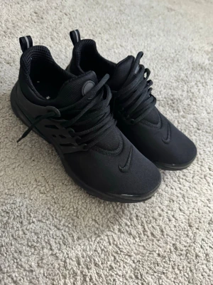 Svarta Nike prestos  - Säljer ett par helsvarta Nike prestos med låg profil och klassisk swoosh på tån. Skorna har mesh- och textilöverdel, svarta skosnören och dragflikar bak. Perfekta för en clean och sportig look.